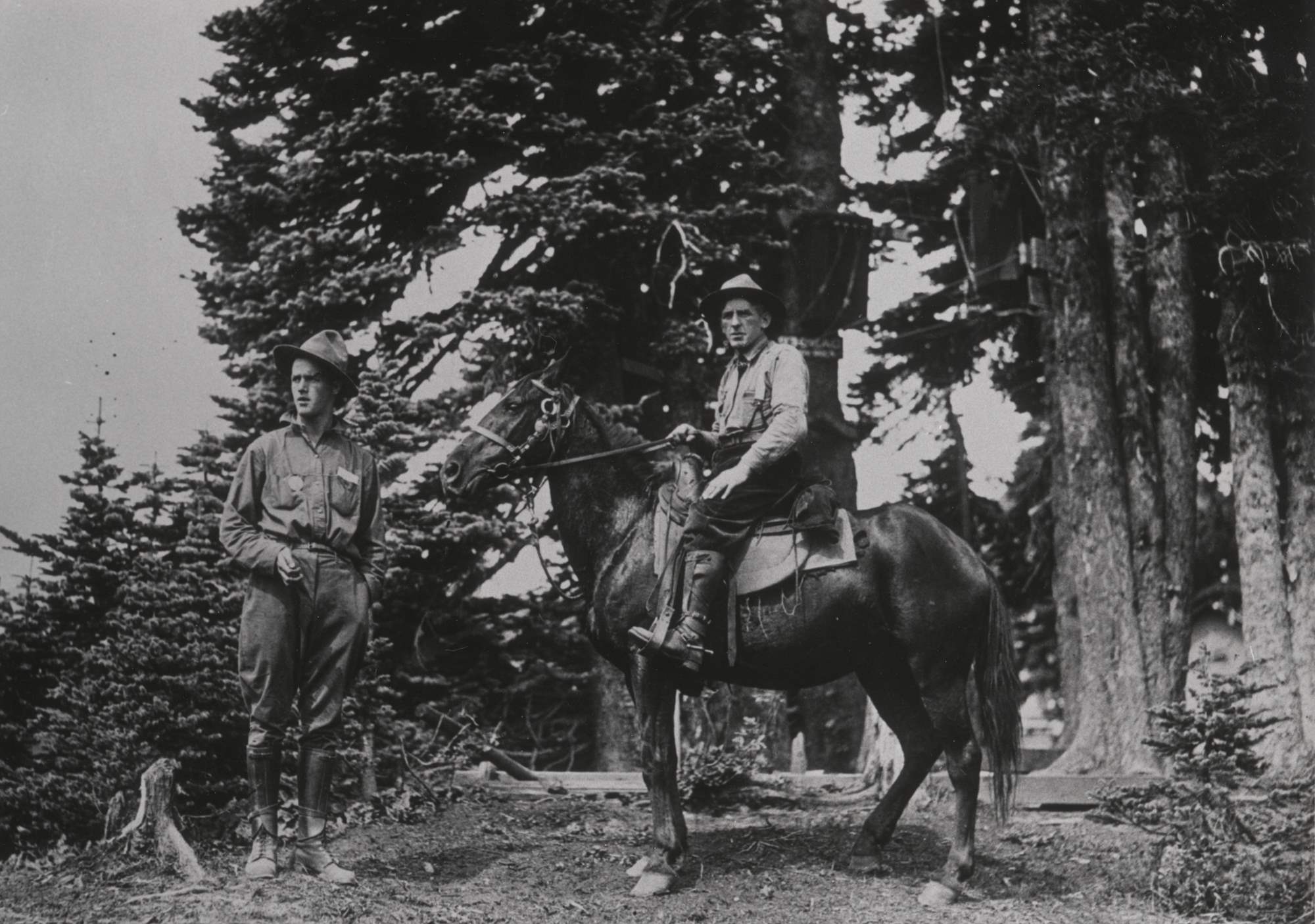 A man stands by another on horseback. Both wear breeches, boots, shirts, and flat brim hats. One has a round badge on his shirt.
